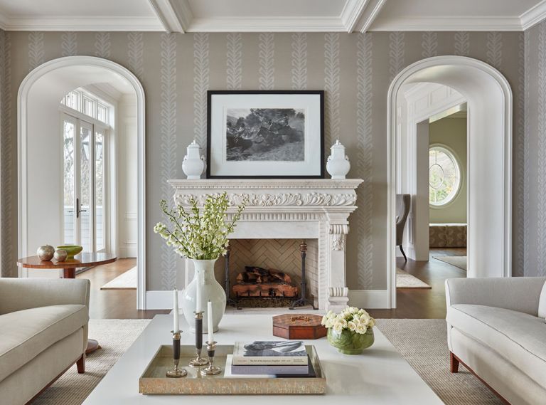 Centered between elliptical arches beneath a traditional boxed ceiling is the cherished, ornate Architectural Supply stone fireplace from Al’s Tile Works framed against Thibaut wallpaper. Custom linen upholstered sofas and a quartz-topped coffee table center the room Centered between elliptical arches beneath a traditional boxed ceiling is the cherished, ornate Architectural Supply stone fireplace from Al’s Tile Works framed against Thibaut wallpaper. Custom linen upholstered sofas and a quartz-topped coffee table center the room