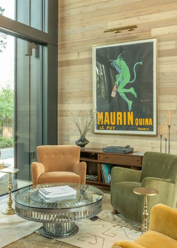 The lounge has a framed 1920s vintage advertisement poster hanging above the custom sideboard. Four Maiden Home armchairs, each in a different color, surround a Timothy Oulton coffee table and all sit atop layered rugs from Lulu and Georgia. The lounge has a framed 1920s vintage advertisement poster hanging above the custom sideboard. Four Maiden Home armchairs, each in a different color, surround a Timothy Oulton coffee table and all sit atop layered rugs from Lulu and Georgia.